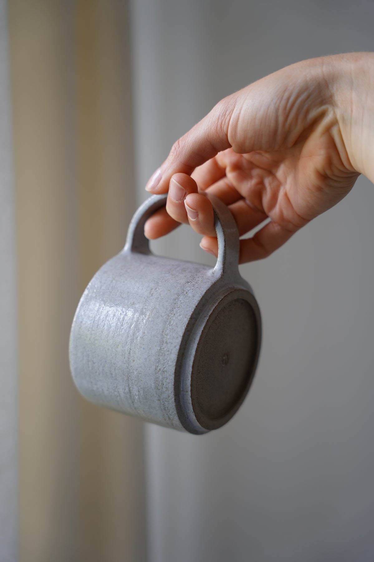 Ein hellgrau, glänzender Keramikbecher aus anthrazit-farbenen Ton mit einem dünnen, halbrund-geformten Henkel, seitlich gehalten, sodass man den unglasierten Boden sieht.