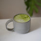 Ein hellgrau, glänzender Keramikbecher aus anthrazit farbenen Ton mit einem dünnen, halbrund-geformten Henkel, gefüllt mit einem leuchtend grünen Matcha-Latte.