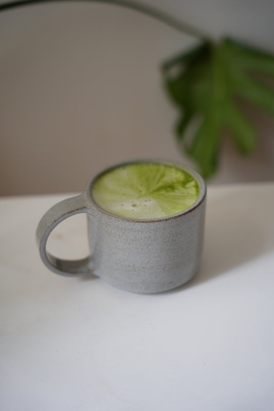 Ein hellgrau, glänzender Keramikbecher aus anthrazit farbenen Ton mit einem dünnen, halbrund-geformten Henkel, gefüllt mit einem leuchtend grünen Matcha-Latte.