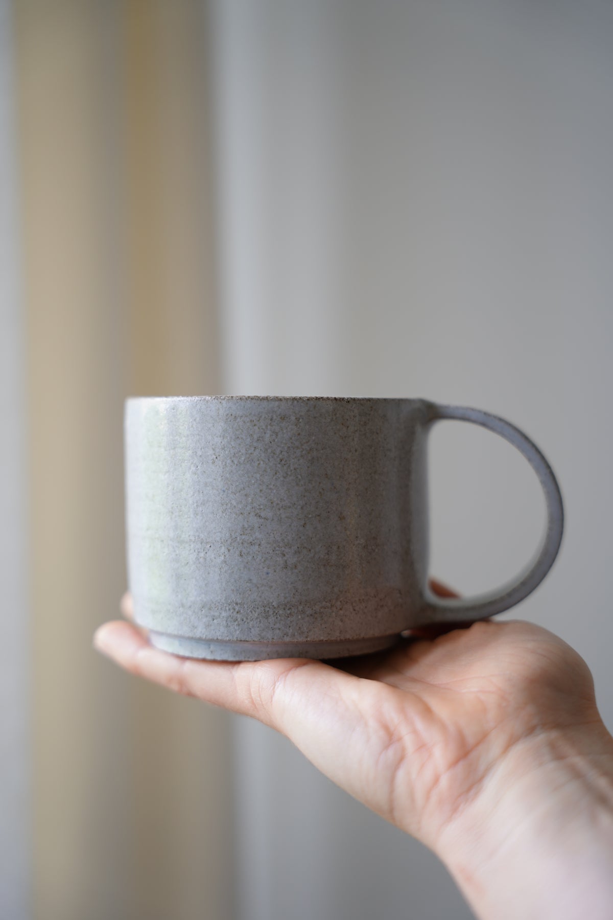 Eine hellgrau, glänzende Keramiktasse aus anthrazit farbenen Ton mit einem dünnen, halbrund-geformten Henkel in der Seitenansicht.