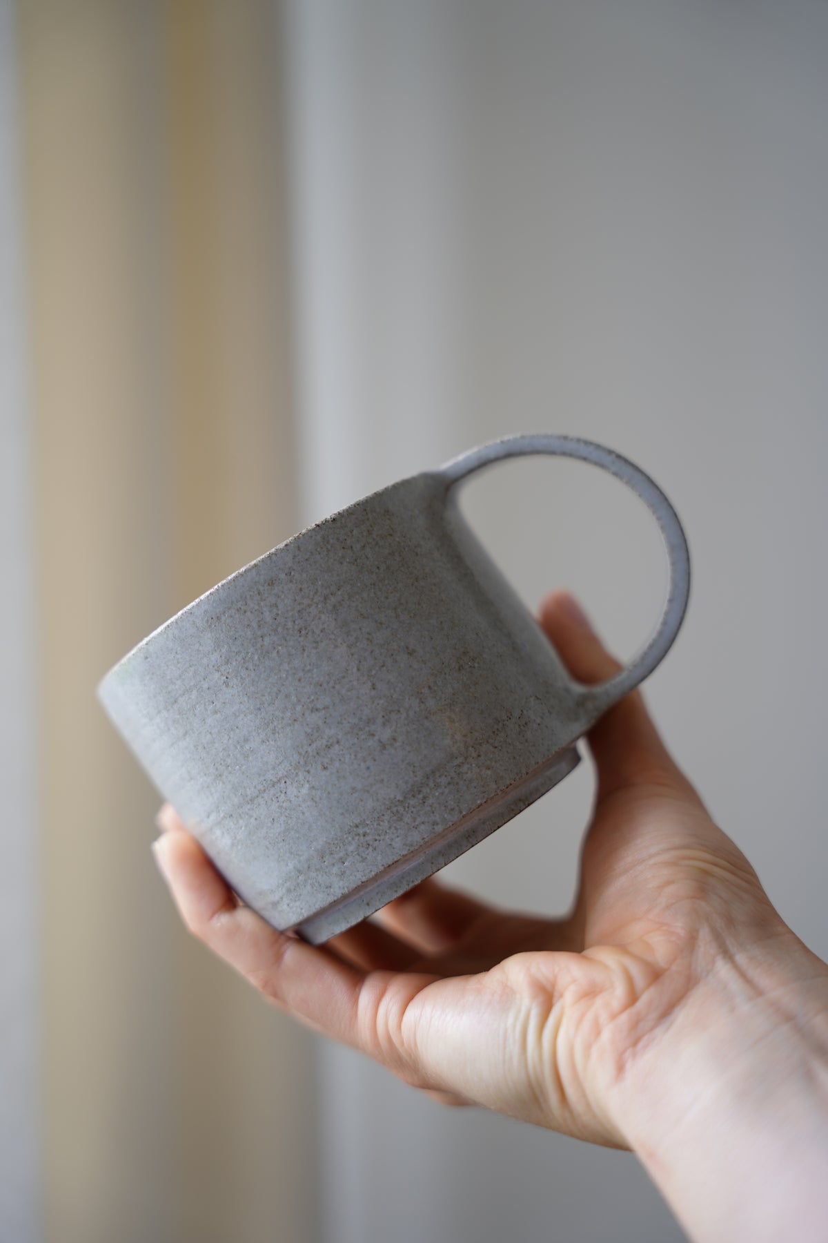 Eine hellgrau, glänzende Keramiktasse aus anthrazit farbenen Ton mit einem dünnen, halbrund-geformten Henkel in der Seitenansicht.